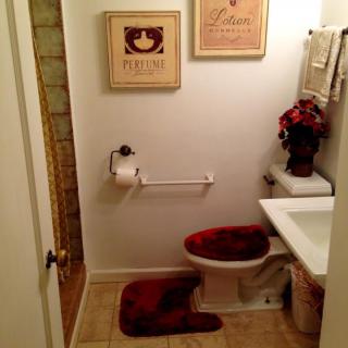 A cozy bathroom with wall art, a toilet adorned with matching red bath mats, and decorative flowers on the sink.