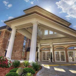 A grand building entrance with large columns, featuring beautiful landscaping and bright sunlight against a clear sky.