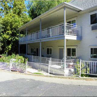 A two-story residential apartment building with balconies and a surrounding fence, set amidst green trees.