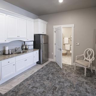 A modern kitchen space with white cabinets and stainless steel appliances, featuring a comfortable chair and access to an adjoining room.