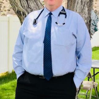 A doctor standing outdoors wearing a shirt, tie, and stethoscope.