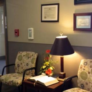 A cozy waiting room with chairs, a lamp, flowers on the table, and certificates on the wall.