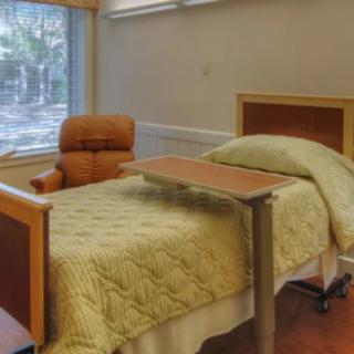 An empty hospital room with a bed, chair, and overbed table near a window.