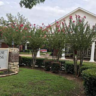A well-maintained building surrounded by lush greenery and flowering trees, with a prominent sign at the entrance.