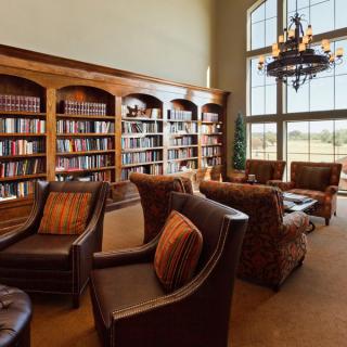 A cozy library space with armchairs and tall bookshelves, illuminated by natural light from a large window.