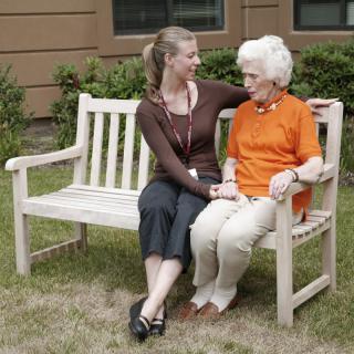 A caregiver and an elderly woman sitting on a bench outdoors, engaged in conversation.