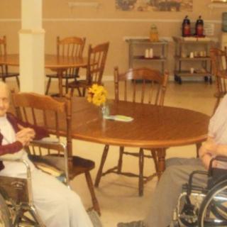 Two elderly individuals in wheelchairs sitting at a table in a nursing home dining area.