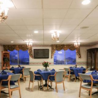 A well-lit dining room with arranged tables and chairs, featuring ceiling lights, a television, and decorated windows.