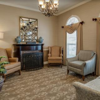 A cozy living room with elegant furnishings, featuring armchairs, a mirror above the mantel, and a chandelier.