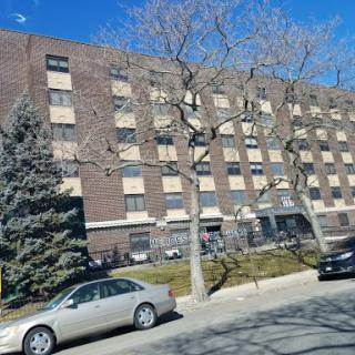 A brick residential building with boarded windows, flanked by trees and parked cars under a clear blue sky.