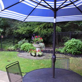 A garden patio with a blue-striped umbrella, surrounded by lush greenery and decorative plants.