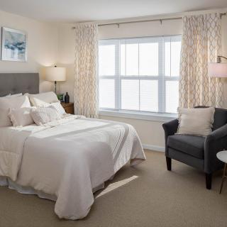 A cozy bedroom with neutral colors, featuring a neatly made bed, a comfortable armchair, and large windows with patterned curtains.
