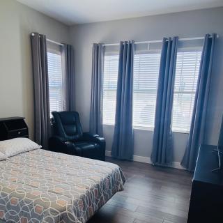 A cozy bedroom featuring a neatly made bed, several large windows with gray curtains, and a comfortable chair.