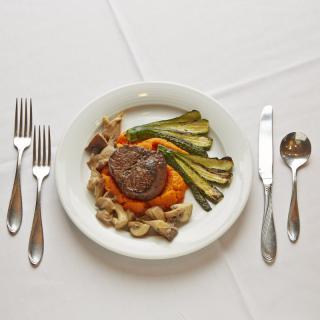 A gourmet meal with steak, vegetables, and pasta served on a white plate with cutlery arranged neatly on a table.