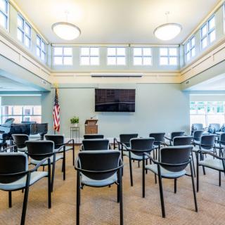 An empty conference room with chairs arranged in rows, featuring a television and podium at the front, alongside an American flag.