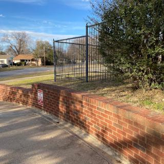 A view of a fenced area next to a brick wall, with grass and a clear blue sky.