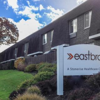 A view of a residential building with a sign for 'eastbro' in the foreground, surrounded by greenery and under a partly cloudy sky.