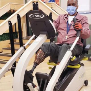 A person using adaptive exercise equipment during rehabilitation, wearing a mask and a prosthetic leg.