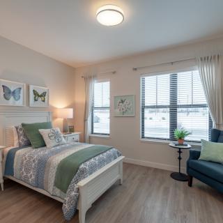 A cozy bedroom with a neatly made bed, artwork on the walls, and a comfortable chair by the window.