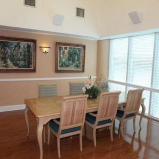 A well-lit dining room with a wooden dining table surrounded by six chairs, adorned with a vase of flowers. Artwork hangs on the wall, and large windows with blinds allow natural light to fill the space.