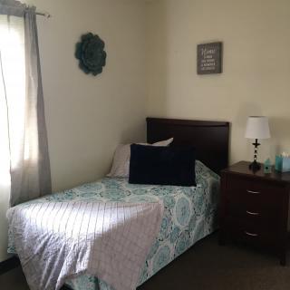 A cozy bedroom featuring a single bed with patterned bedding, a nightstand with a lamp, and wall decor.
