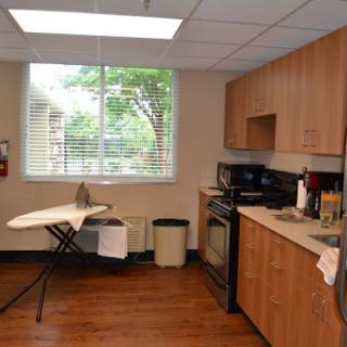 A cozy kitchen space featuring wooden cabinets, appliances, and an ironing board set up by the window.