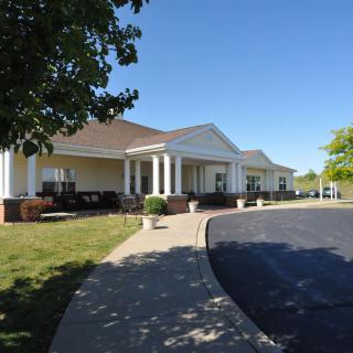 A large building with a columned entrance and a driveway, set in a landscaped area on a sunny day.