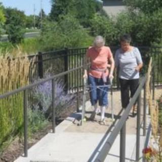 An elderly person with a walker is assisted by a caregiver on an outdoor path.