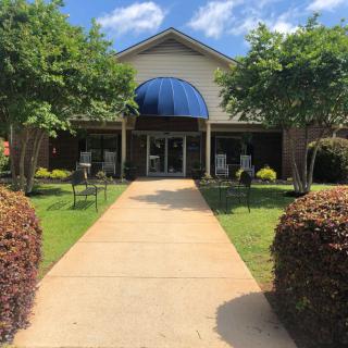 A walkway leads to a building entrance with a blue awning, surrounded by trees and neatly trimmed bushes.