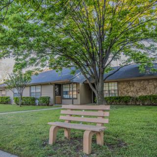 A suburban scene featuring a bench on a lush green lawn in front of a house with trees.