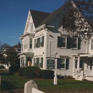 A large white residential house with green shutters and a well-maintained lawn on a sunny day.