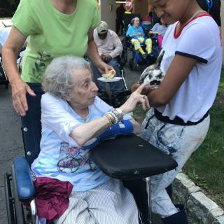An elderly woman in a wheelchair interacts with a young person holding a small dog outdoors.