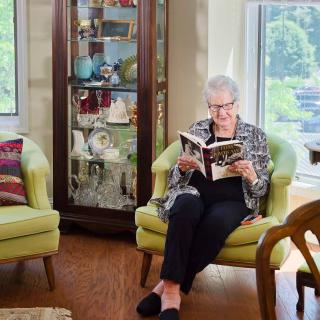 An elderly woman sits comfortably in an armchair, reading a book in a cozy living room.