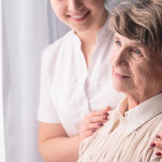 A caregiver providing support and companionship to an elderly woman near a window.