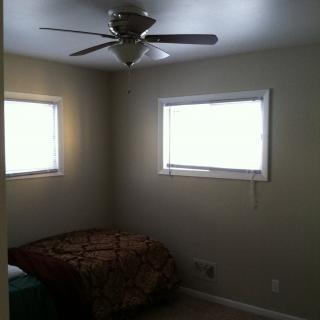 A minimalist bedroom featuring a ceiling fan, two windows, and a simple bed.