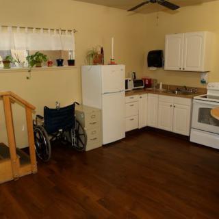 An accessible kitchen with a wheelchair, featuring white cabinets, appliances, and a wooden floor.