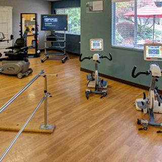 A rehabilitation therapy room equipped with exercise bikes and parallel bars on a wooden floor.