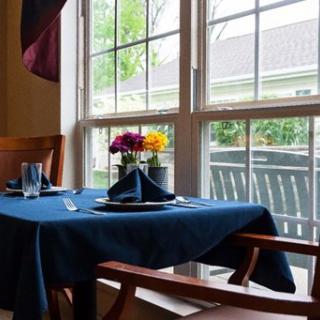A cozy dining area with a table set for two, featuring flowers and a view of the outside through the window.