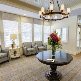 A well-lit lobby with comfortable seating, featuring a central round table adorned with a floral arrangement and an elegant chandelier overhead.