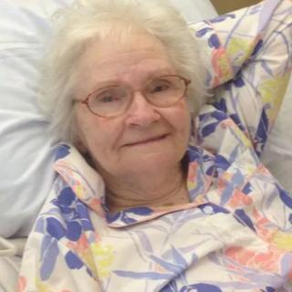 An elderly woman with glasses smiling while lying in a hospital bed.