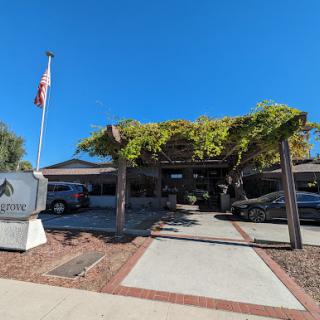 The entrance of a building under a clear blue sky, featuring a flag and lush greenery.