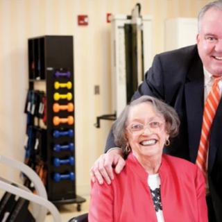 An elderly woman smiling with a man in a suit beside her, in a room with fitness equipment.
