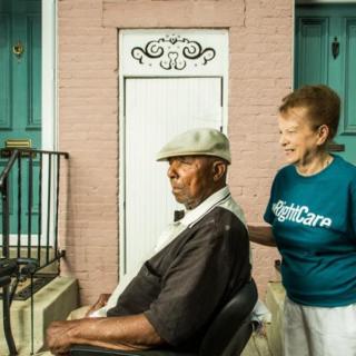 A caregiver assisting an elderly man in a wheelchair outside a building.