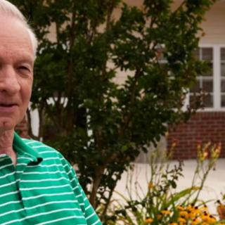 An elderly man in a green striped shirt stands outside near a garden.