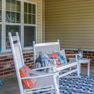 A cozy porch setup with white rocking chairs and a bench, decorated with colorful pillows and lanterns on a patterned rug.