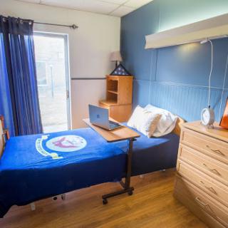 A cozy bedroom featuring a neatly made bed, a laptop on a small desk, and warm wooden furniture.
