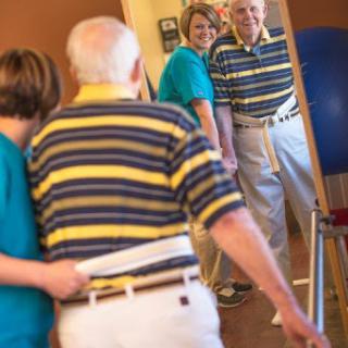 An elderly man receives support during a rehabilitation session, as he practices standing and walking in front of a mirror.