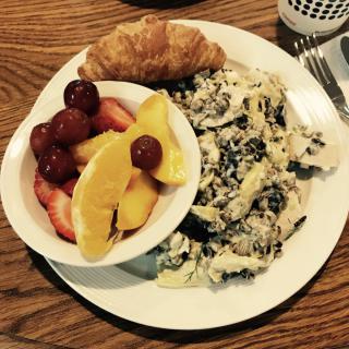A hearty breakfast featuring a croissant, scrambled eggs, and a bowl of fresh fruit including grapes, strawberries, and orange slices.