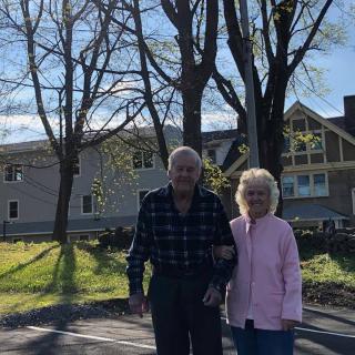 An elderly couple walking outdoors near a house with trees in the background.
