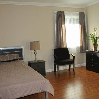 A minimalist bedroom with simple furnishings, including a bed, chair, lamp, and dresser.